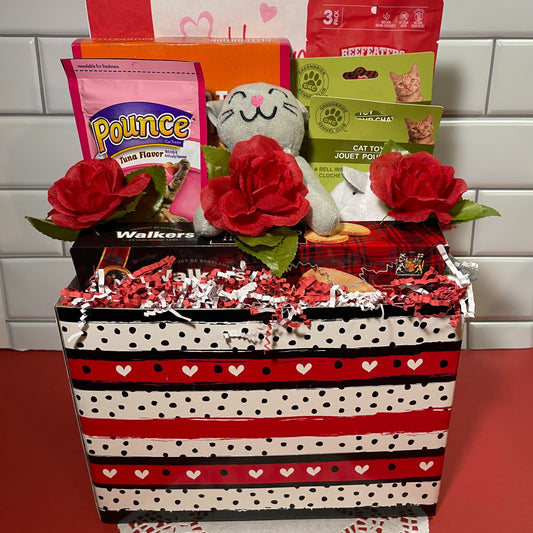 a red, white and black striped box filled with a plush gray cat, a red bird cat toy, a stuffed mouse cat toy, a package of puree cat treats, a package of heart-shaped dry cat treats, a box of Walker's shortbread cookies, a box of raspberry truffle butter cookies, 3 red roses and a Valentine's Day napkin. The box is sitting on a white doily, in front of a white background.