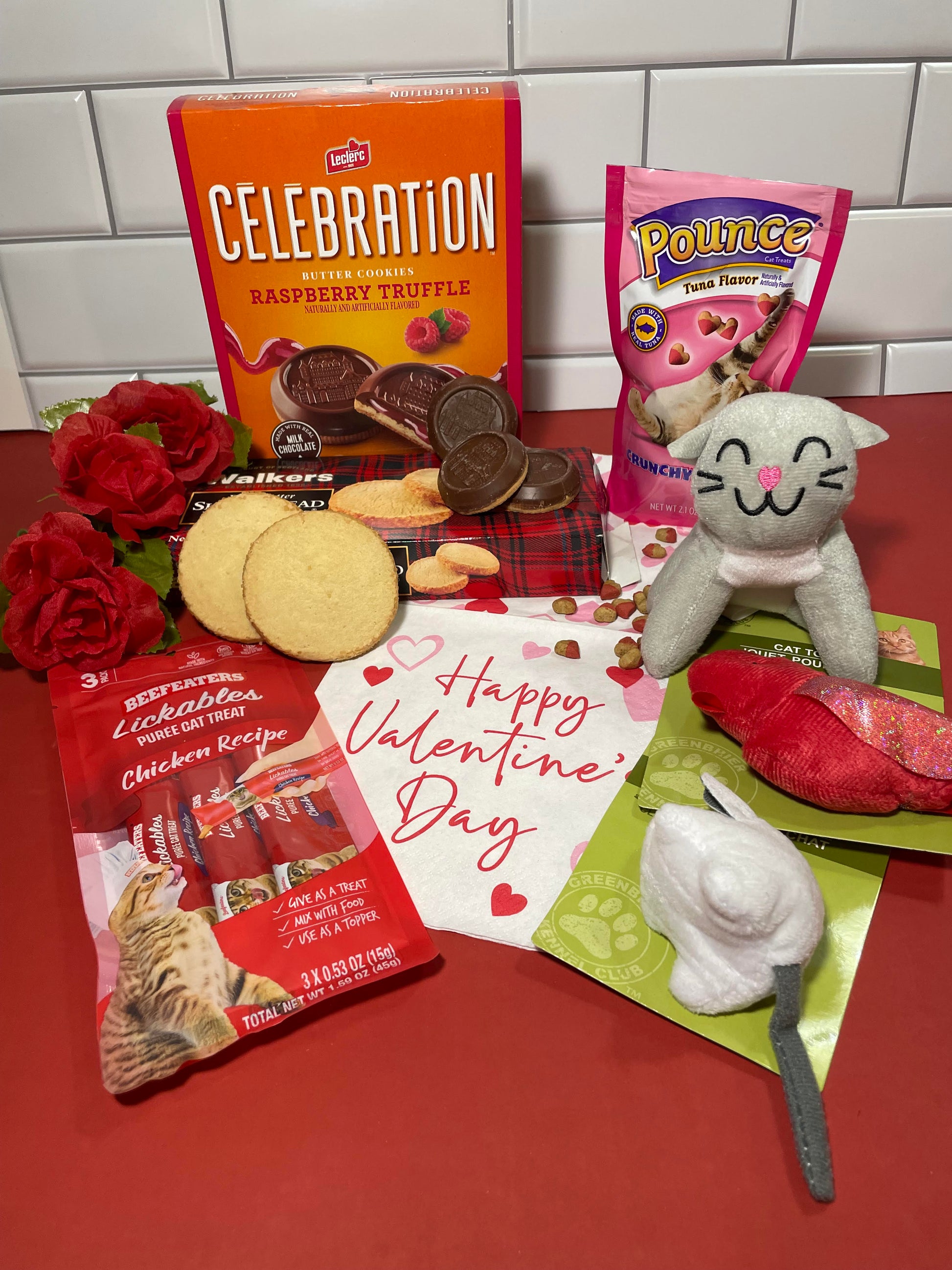 3 red roses, a box of raspberry truffle cookies, a box of Walker's Shortbread cookies, two cat toys. a grey stuffed cat, a package of dry heart-shaped cat treats and a package of 3 pueed cat treats all on a Happy Valentine's Day napkin