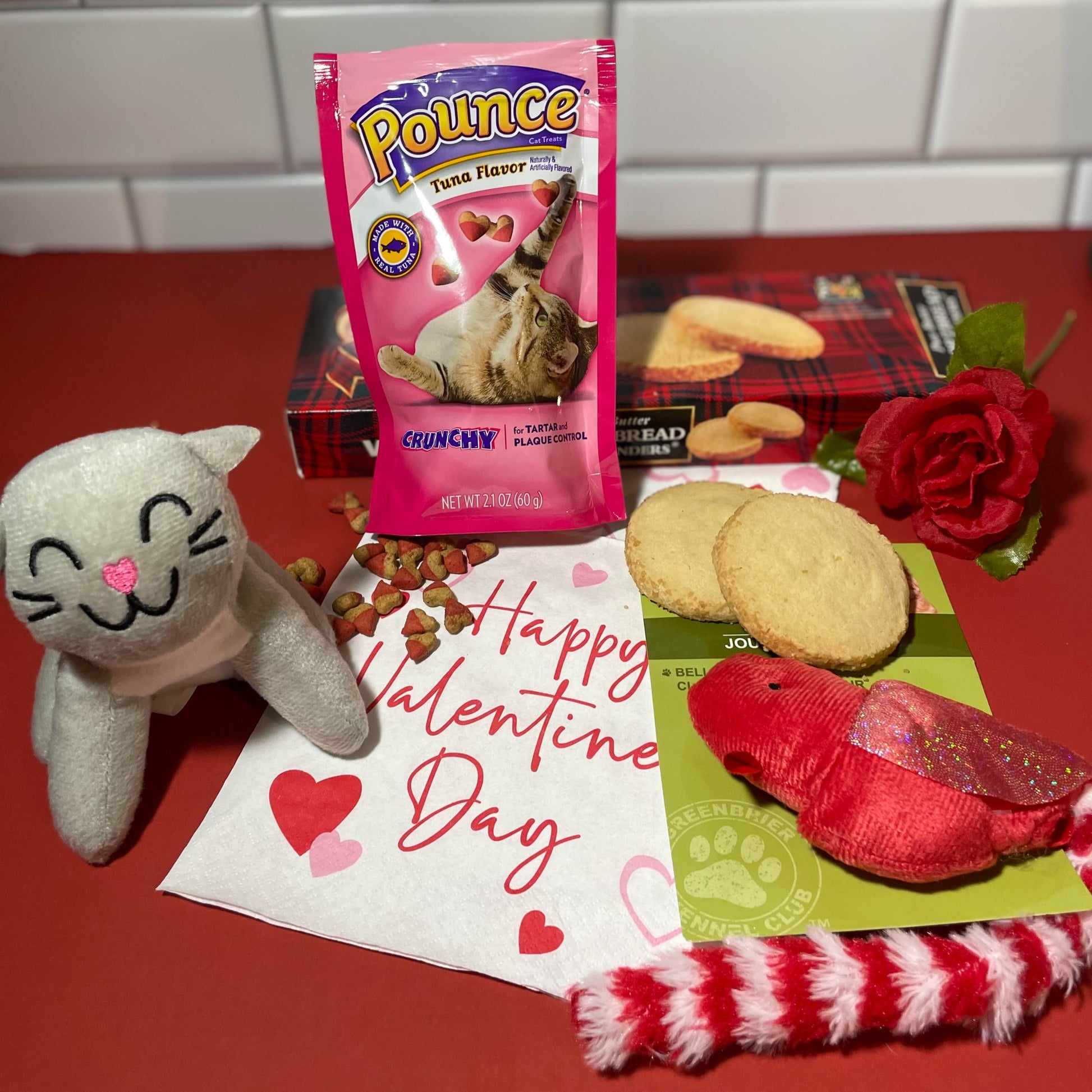 a grey stuffed cat, pouch of heart-shaped cat treats, red cat toy shaped like a bird. a red silk rose and a box of Walker's shortbread cookies all arranged on a Happy Valentine's napkin