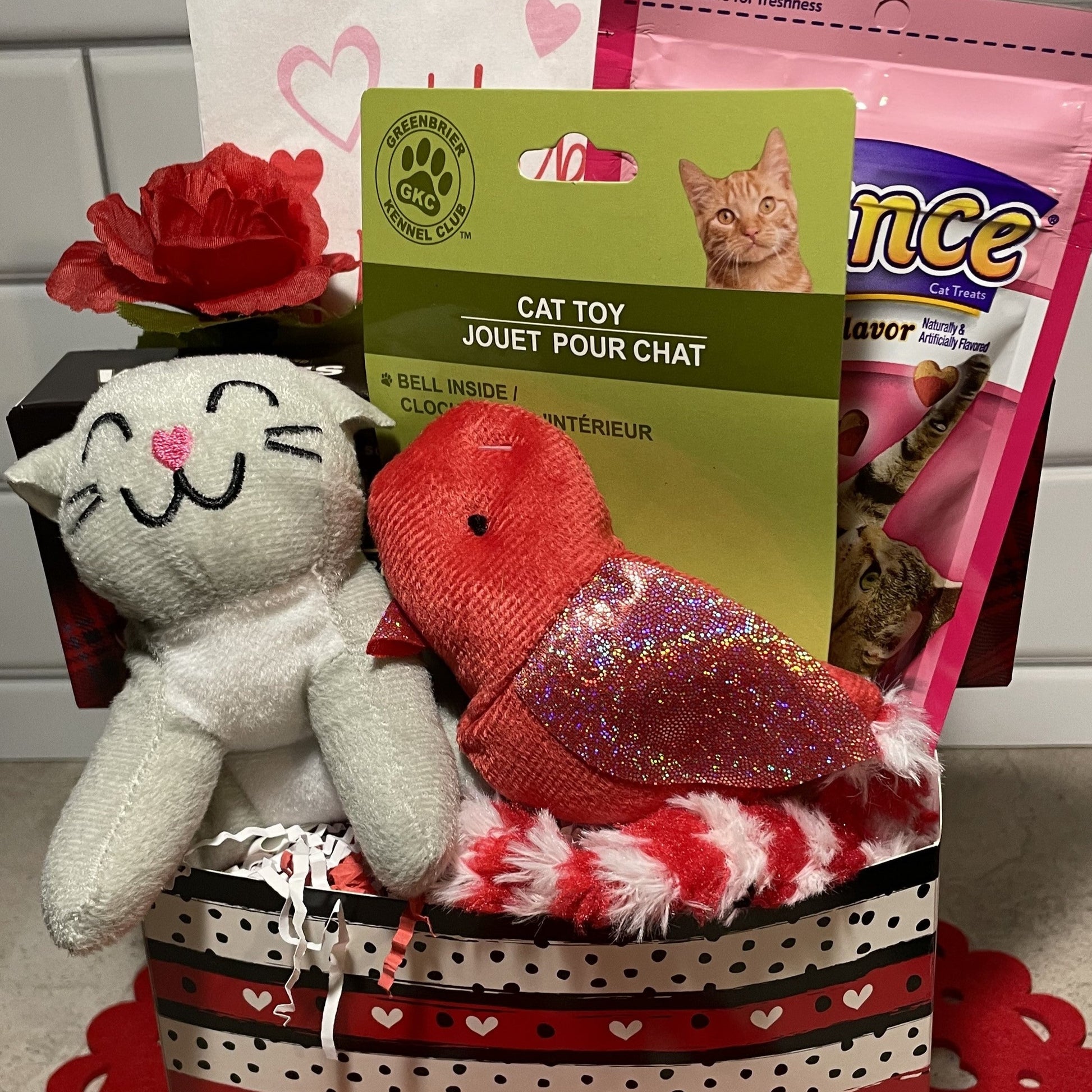 a red, white and black striped box filled with a plush gray cat, a red bird cat toy, a package of cat treats, a box of Walker's shortbread cookies, a red rose and a Valentine's Day napkin. The box is sitting on a red lace heart, in front of a white background. 